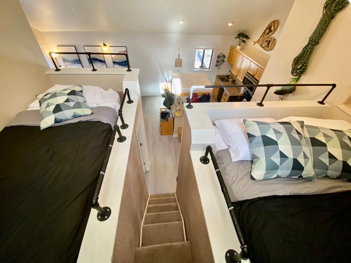 This image features a bright interior with lofted beds visible above the kitchen area, a staircase leading up to the lofts, and inviting decorative elements on the walls. Natural light streams through the windows, creating a welcoming and airy atmosphere. The setup clearly highlights the unique, cozy sleeping nooks elevated beneath high ceilings.