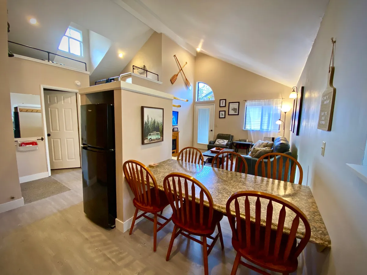 A warm and welcoming open-concept space that encompasses a dining table, kitchen area, and living room seating, all framed by large windows. The space is filled with natural light, further enhanced by the view to the outside, which subtly suggests the presence of a balcony or patio area beyond.