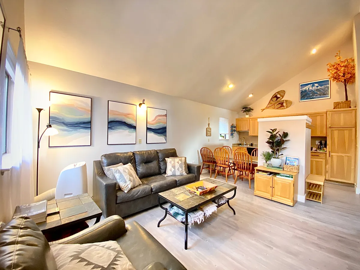 A welcoming open-plan interior featuring a comfortable sofa with plush cushions, a dining table and chairs set for gatherings, and tasteful artwork and decorative plants that create a homey atmosphere. The layout suggests a space where families can relax, enjoy meals, and play games together.