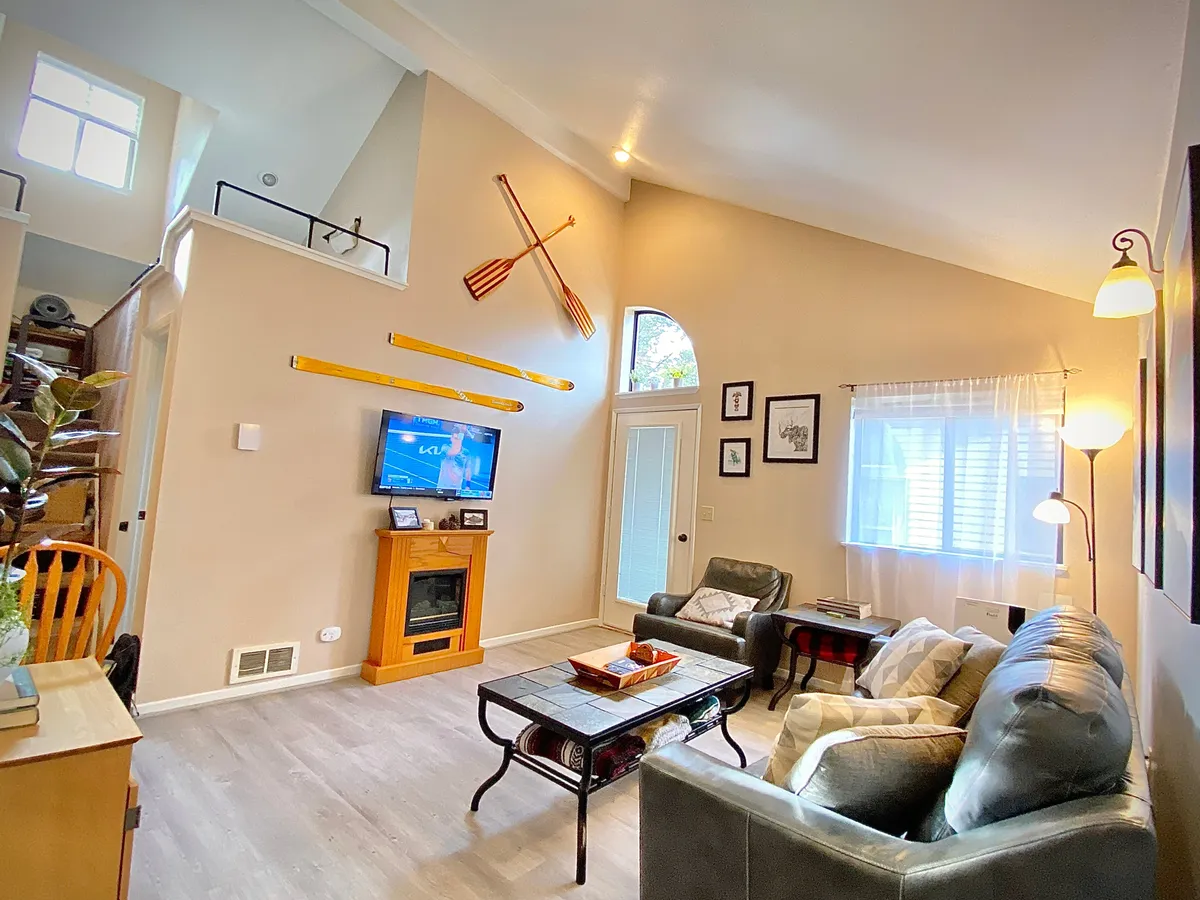 This image features a cozy living area with comfortable seating arranged to face a TV mounted above a fireplace. Large windows dominate one wall, letting in natural light and offering glimpses of the outdoors. Decorative oars and homey touches contribute to a resort-like, inviting atmosphere. The view through the windows hints at the connection between the indoor space and the balcony just outside.