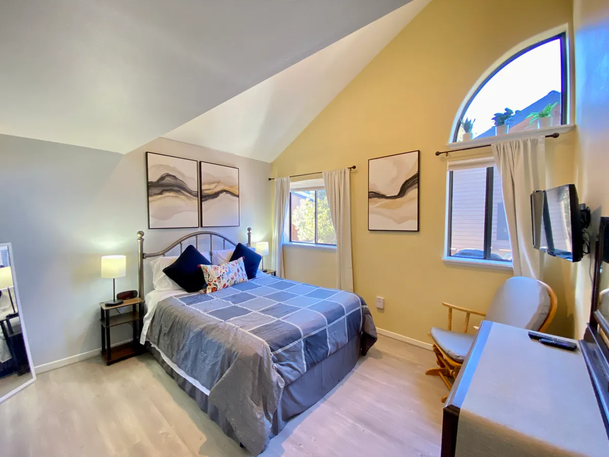 A serene bedroom setting with a queen bed adorned with decorative pillows, understated wall art, and abundant natural light pouring in through a large window. The room feels peaceful and comfortable, ideal for relaxation and restorative sleep.