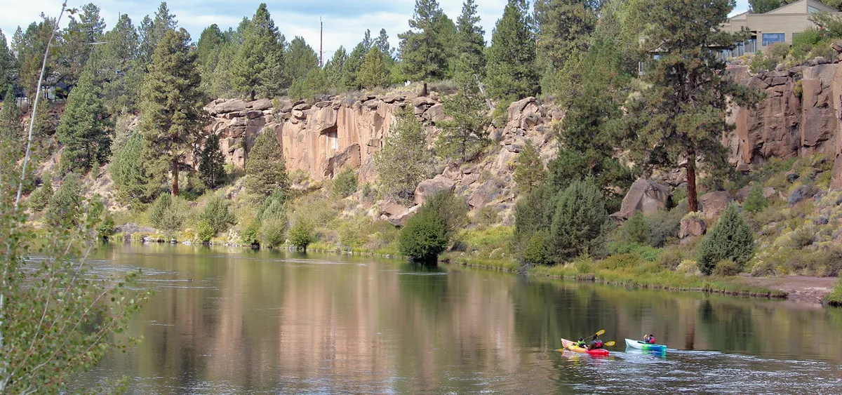A scenic view of the Deschutes River framed by rocky cliffs and lush trees, with kayakers paddling along the water. The natural surroundings showcase the tranquility and adventure opportunities just moments away from the property.