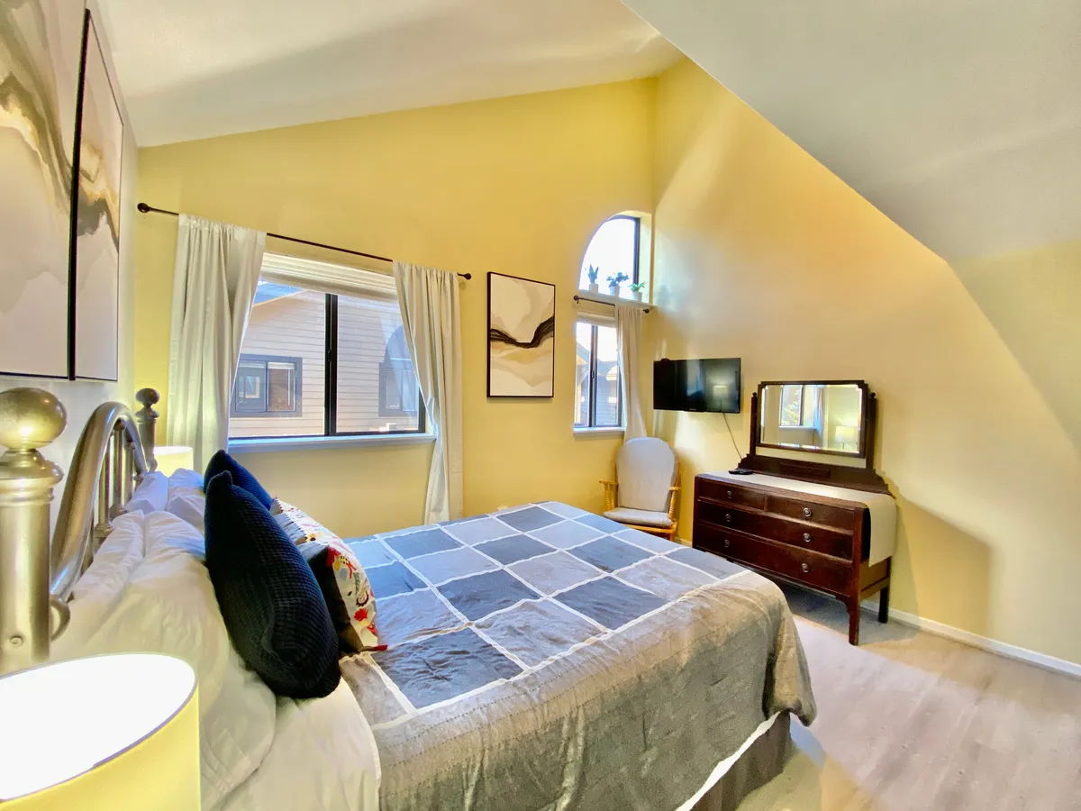 A cheerful bedroom with a queen-sized bed dressed in patterned bedding, large sunny windows, a wooden dresser, and colorful artwork. The space is cozy and bright, offering a restful retreat with a welcoming vibe.