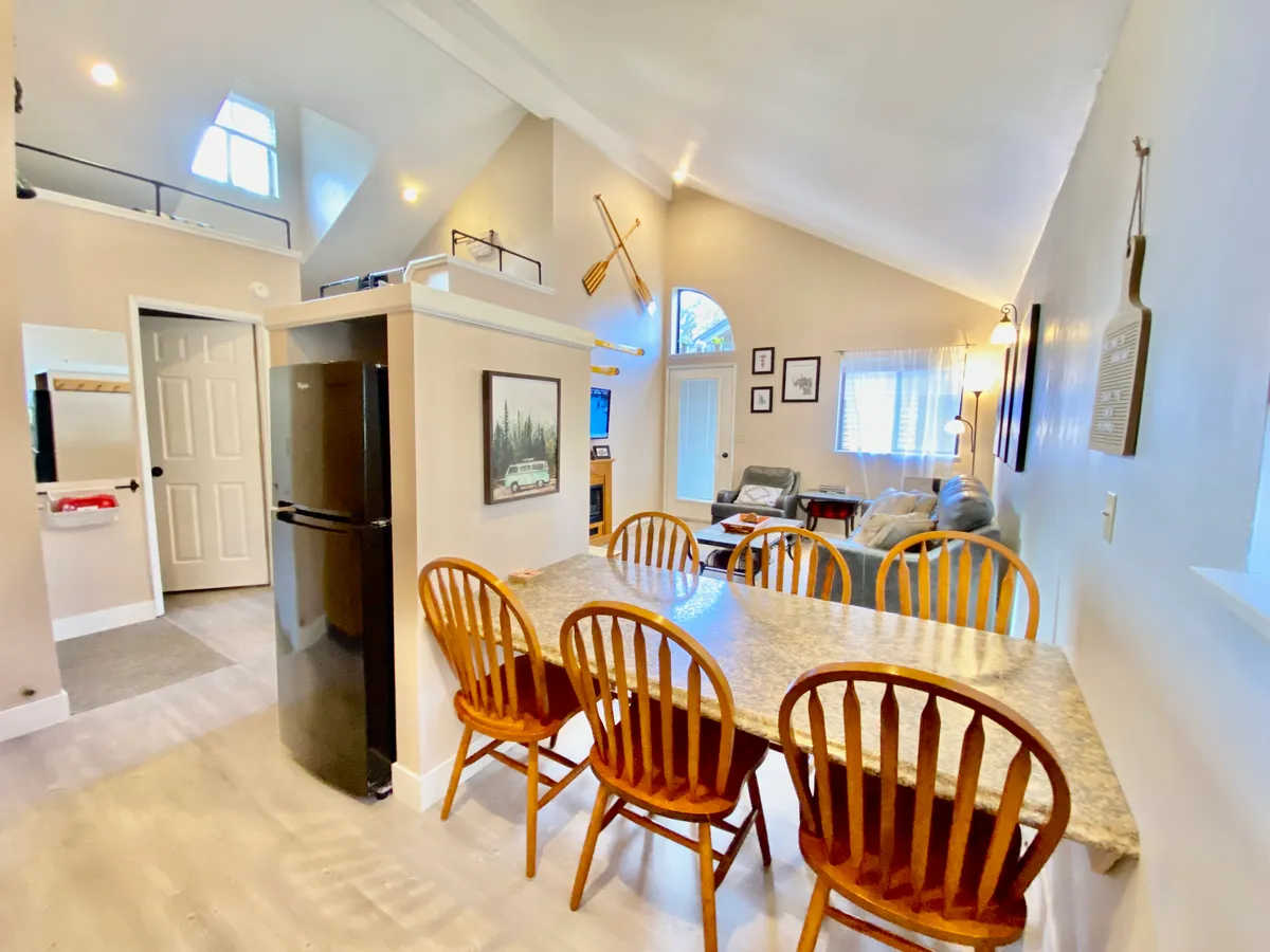 A spacious, sunlit open-concept area featuring a dining table set for family meals, a fridge in the background, comfortable living area furnishings, and expansive windows that flood the room with natural light. Decorative touches and board games invite social gatherings and relaxation.